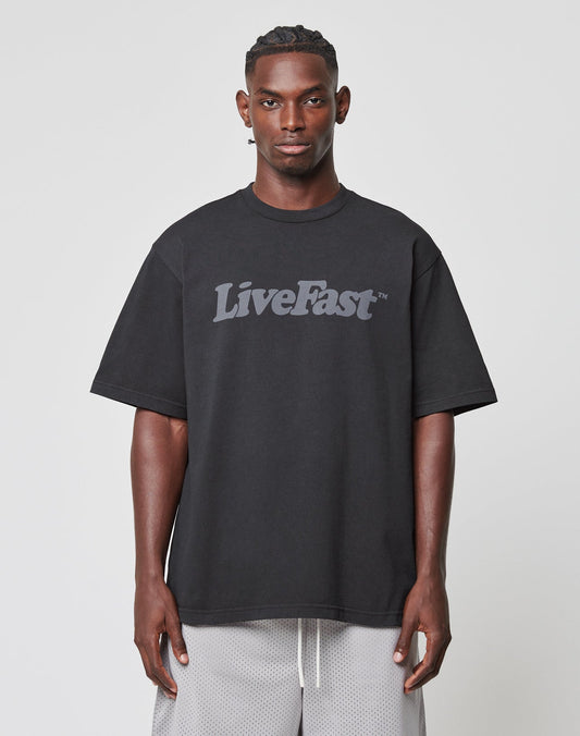 A man with short, styled hair wears the LIVE FAST DIE YOUNG Basic Tee in an oversize fit, featuring bold gray Live Fast text on 100% cotton, paired with light gray mesh shorts against a plain background.