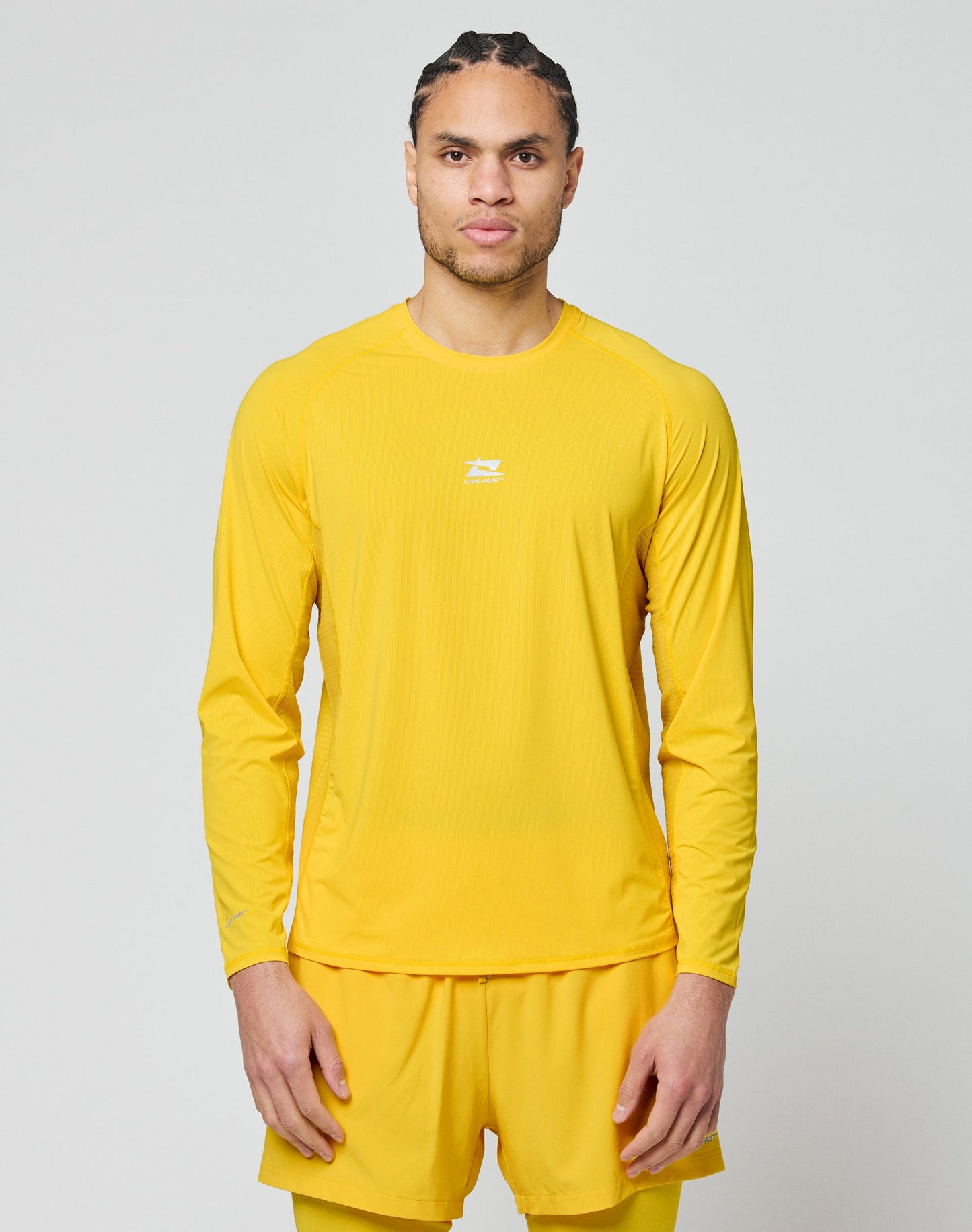 A person with braided hair stands against a plain light gray background, wearing the LIVE FAST DIE YOUNG Live Fast Performance Longsleeve in yellow and matching shorts, looking straight ahead with a neutral expression.