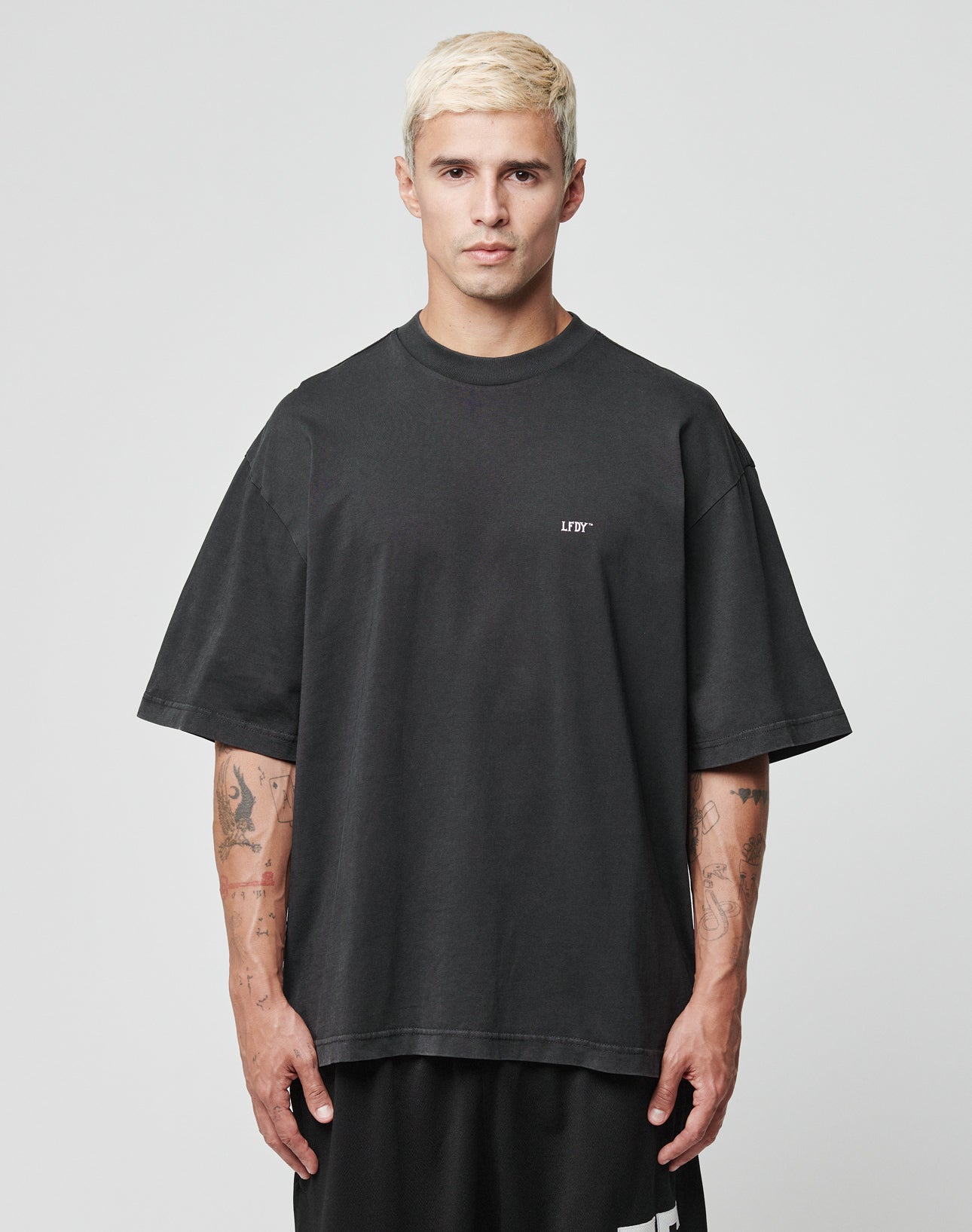 A man with short bleached blonde hair wears the LIVE FAST DIE YOUNG Everybody Eats Tee, an oversize black 100% Baumwolle T-shirt, featuring logo on the chest. He stands facing forward against a plain light background, arms relaxed to show tattoos.