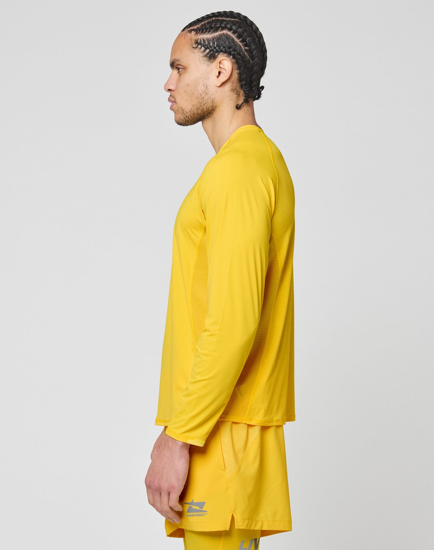 A man with braided hair is shown in profile, wearing the LIVE FAST DIE YOUNG Live Fast Performance Longsleeve and matching yellow shorts, standing against a plain light gray background.