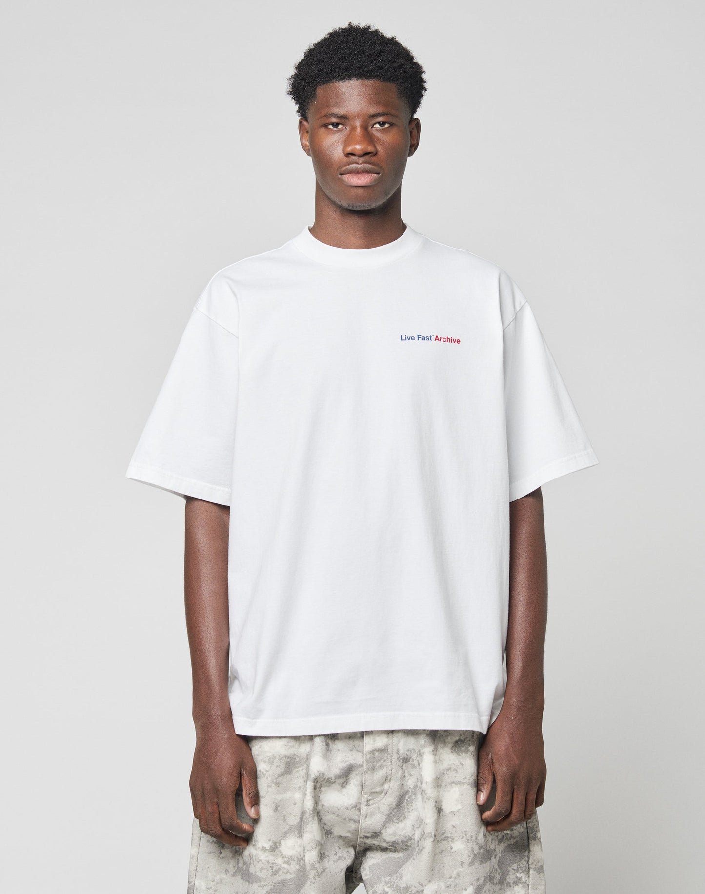 A man stands facing forward wearing the LIVE FAST DIE YOUNG LFDY F*ck Phones Tee, a 100% Baumwolle plain white Oversize Fit T-shirt with small red text on the upper chest, and light gray camo-patterned pants against a light gray background.