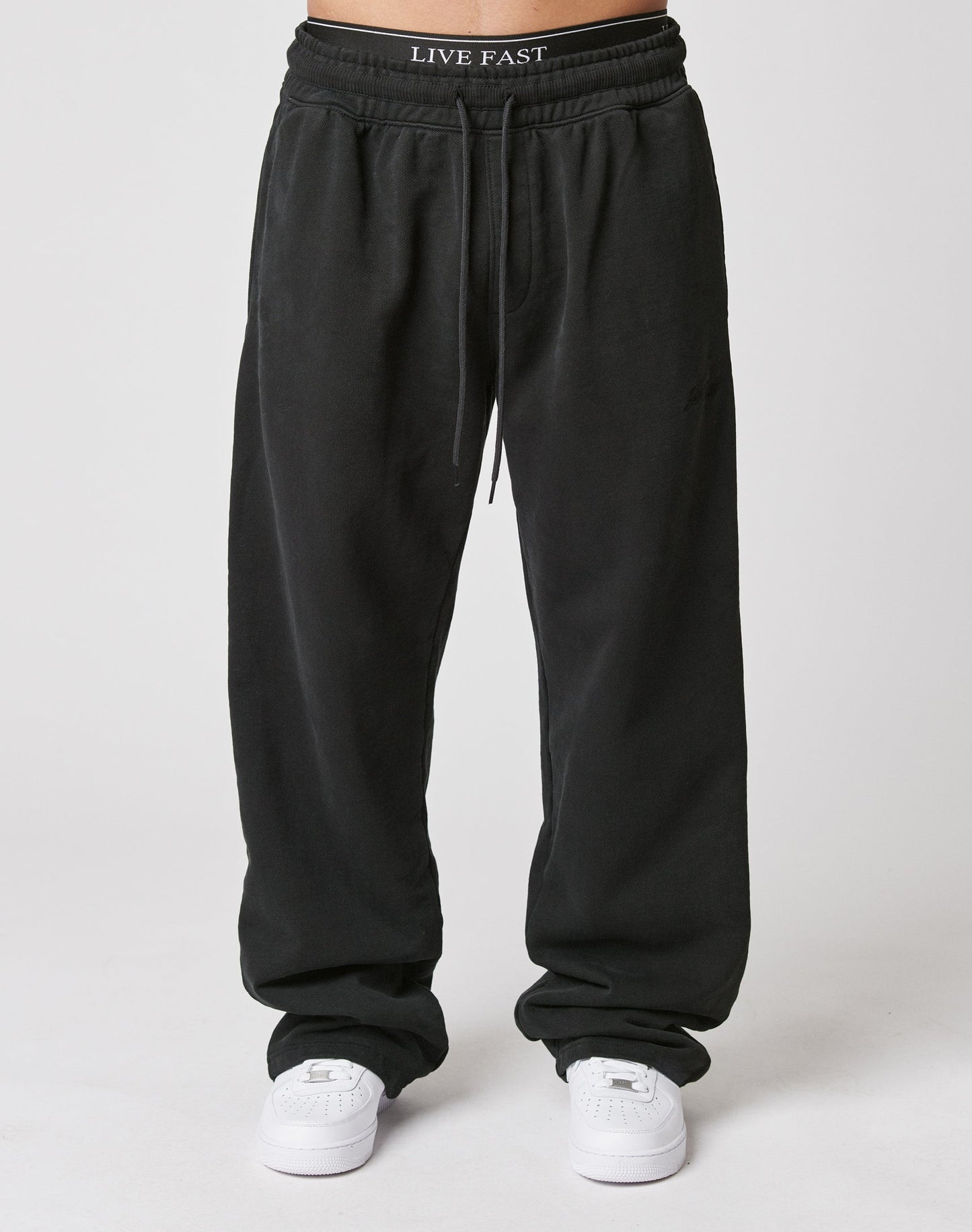 A person wears LIVE FAST DIE YOUNG’s Basic Wide Jogger in black with an elastic waistband, paired with white sneakers and LIVE FAST waistband underwear, standing against a plain white background.