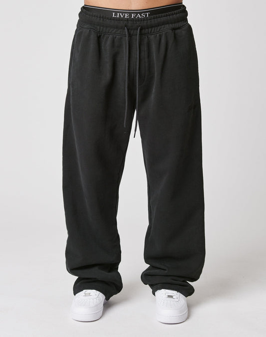 A person wears LIVE FAST DIE YOUNG’s Basic Wide Jogger in black with an elastic waistband, paired with white sneakers and LIVE FAST waistband underwear, standing against a plain white background.