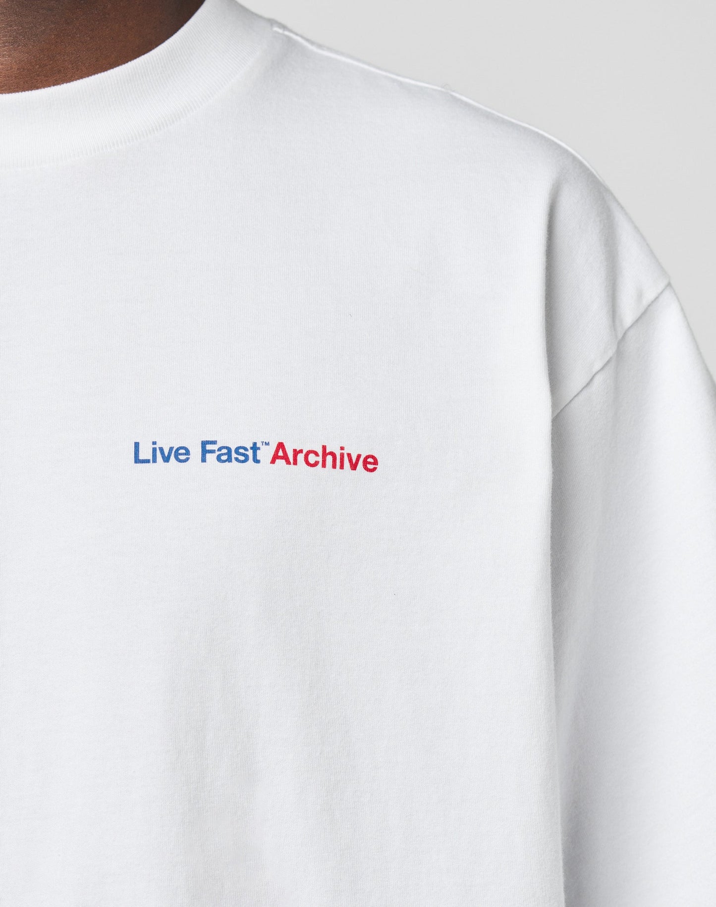 Close-up of a person wearing the LFDY F*ck Phones Tee by LIVE FAST DIE YOUNG—an oversized white cotton T-shirt with “Live Fast Archive” in blue and red on the chest. Only the upper torso is visible; features both front and back print designs.