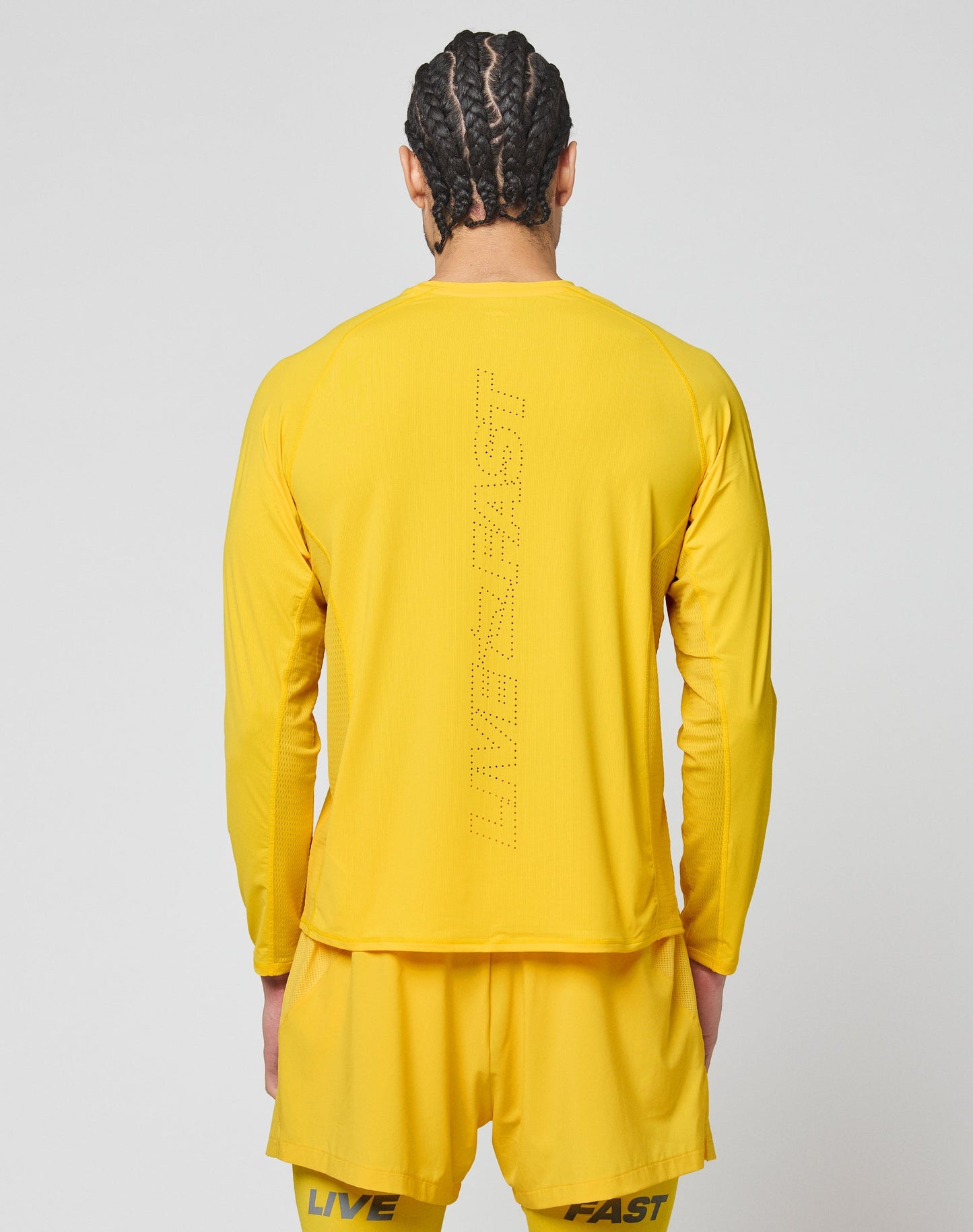 A person with braided hair, seen from behind, wears a bright yellow LIVE FAST DIE YOUNG Live Fast Performance Longsleeve and matching shorts, both featuring LIVE FAST printed on the back.
