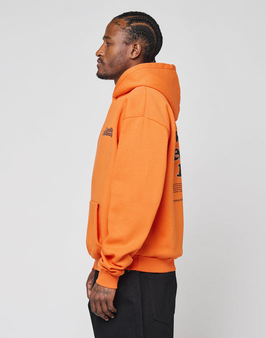 A person with braided hair stands in profile wearing the LIVE FAST DIE YOUNG Basic Orange Hooded sweatshirt with black front and back prints, and black pants, against a plain light gray background.