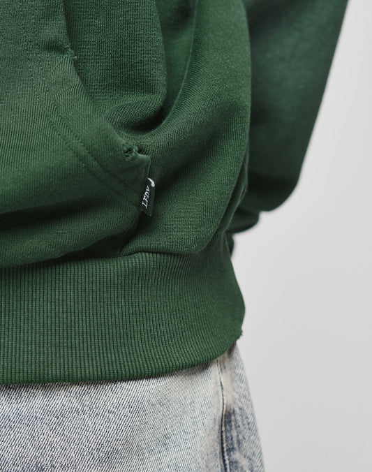Close-up of a person in the LIVE FAST DIE YOUNG LFDY Script Hooded, featuring an Oversize Fit in dark green with a small black tag near the hem, paired with light blue denim jeans. The plain background highlights its modern style.