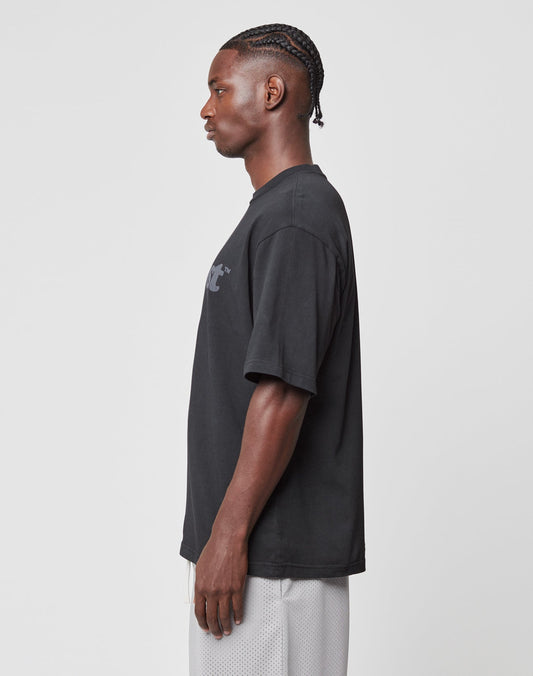 A man stands in profile against a plain background, wearing the LIVE FAST DIE YOUNG Basic Tee in black with a small chest graphic and light gray shorts. His braided hair and relaxed pose enhance the casual vibe.