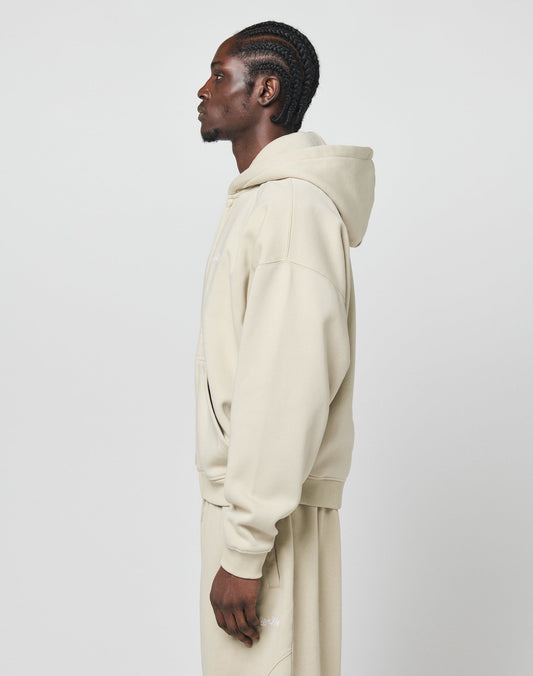 A person with braided hair stands in profile, wearing the LIVE FAST DIE YOUNG Basic Script Hooded Zip in beige and matching sweatpants made from soft Baumwolle against a plain light background.