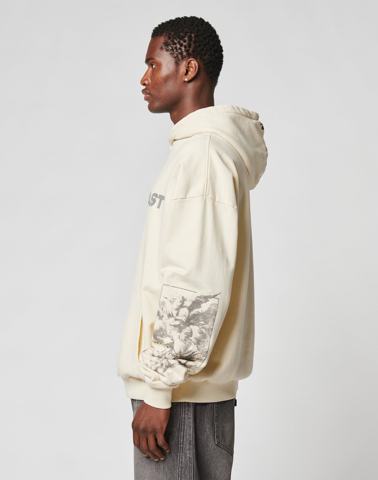 Someone in profile stands against a light background wearing the LIVE FAST DIE YOUNG LFDY Window Hooded, an oversize light beige hoodie with reflektierender Druck, a graphic print on the left sleeve, front pocket, and paired with gray jeans.