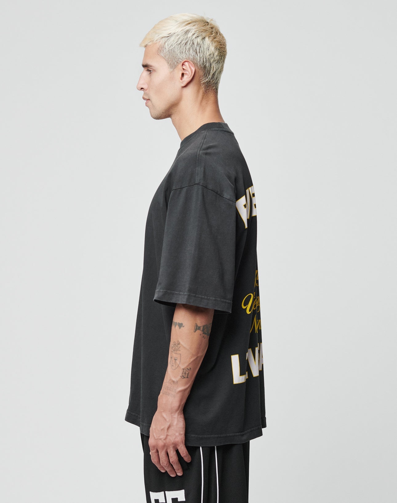 A man with short blond hair wears the LIVE FAST DIE YOUNG Everybody Eats Tee, an Oversize Fit black T-shirt with yellow and white text on the back, paired with black athletic shorts, standing in profile against a plain light gray background.
