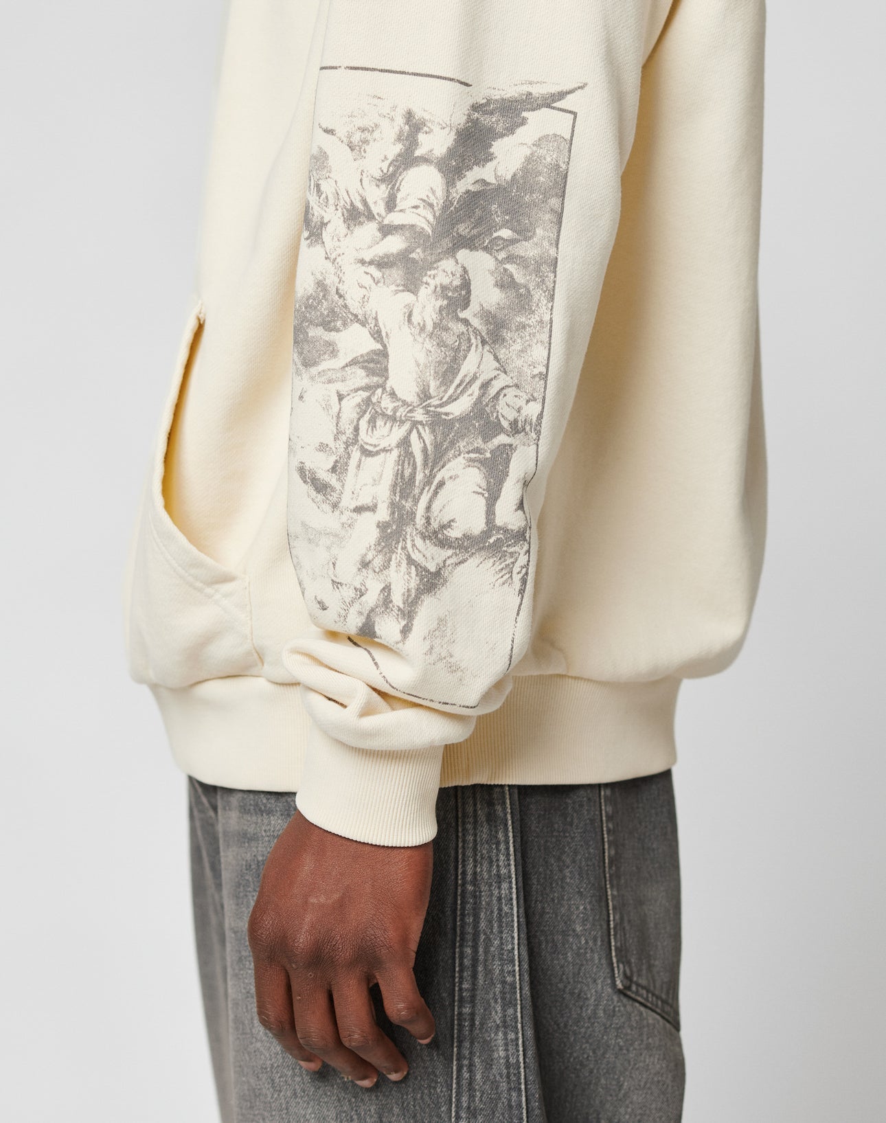 A person wearing the LIVE FAST DIE YOUNG LFDY Window Hooded sweatshirt in cream with a black-and-white classical art print and reflective sleeve print, paired with gray jeans. The image shows their torso and arm from the side.
