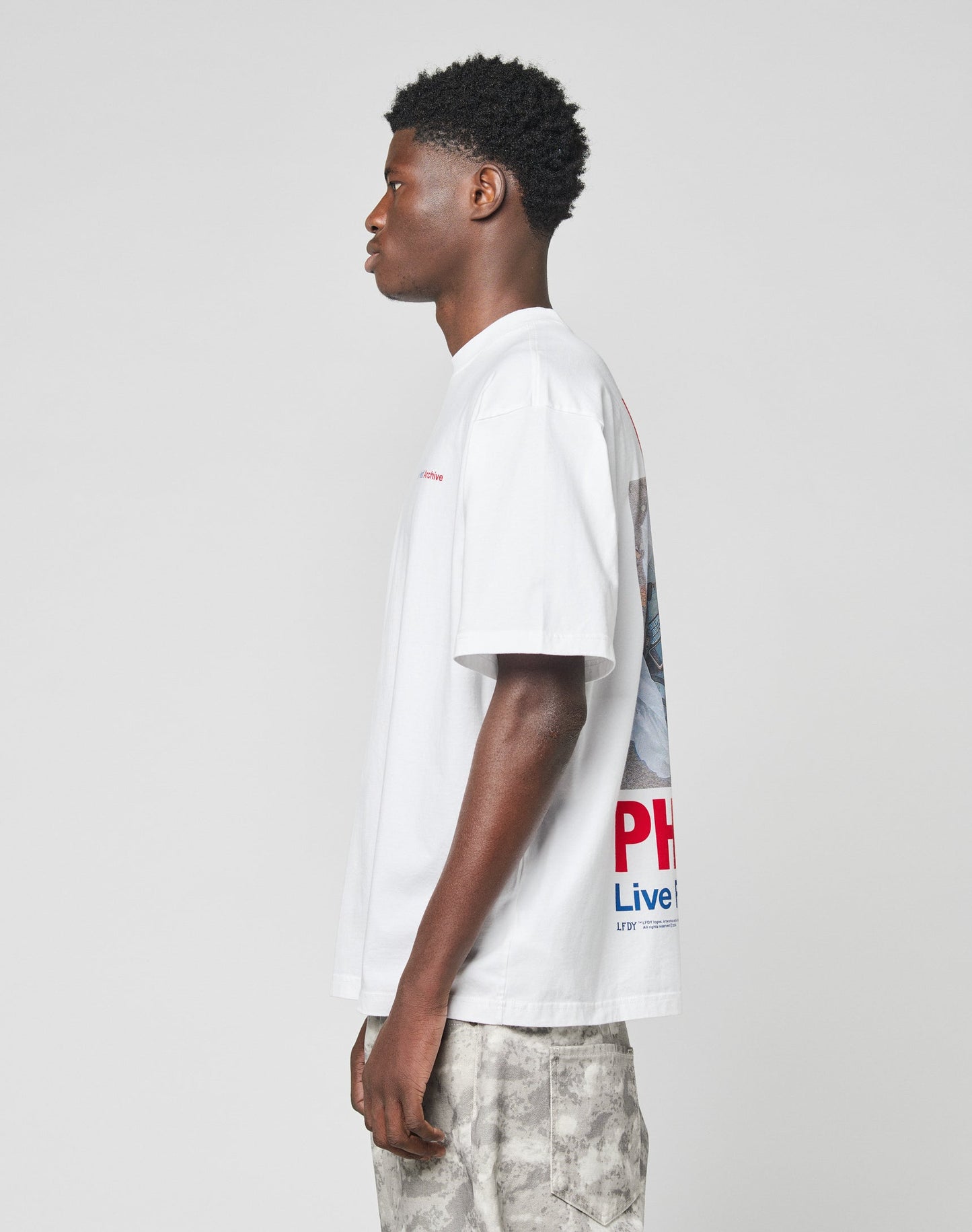 A young man stands in profile against a plain background, wearing the LIVE FAST DIE YOUNG LFDY F*ck Phones Tee in oversize fit with colorful front and back print, paired with light gray tie-dye shorts.