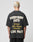 A person with short, bleached blond hair wears the LIVE FAST DIE YOUNG Everybody Eats Tee, a black Oversize Fit shirt made of 100% Baumwolle with bold yellow and white text on the back. Plain gray background; care label includes waschhinweise.