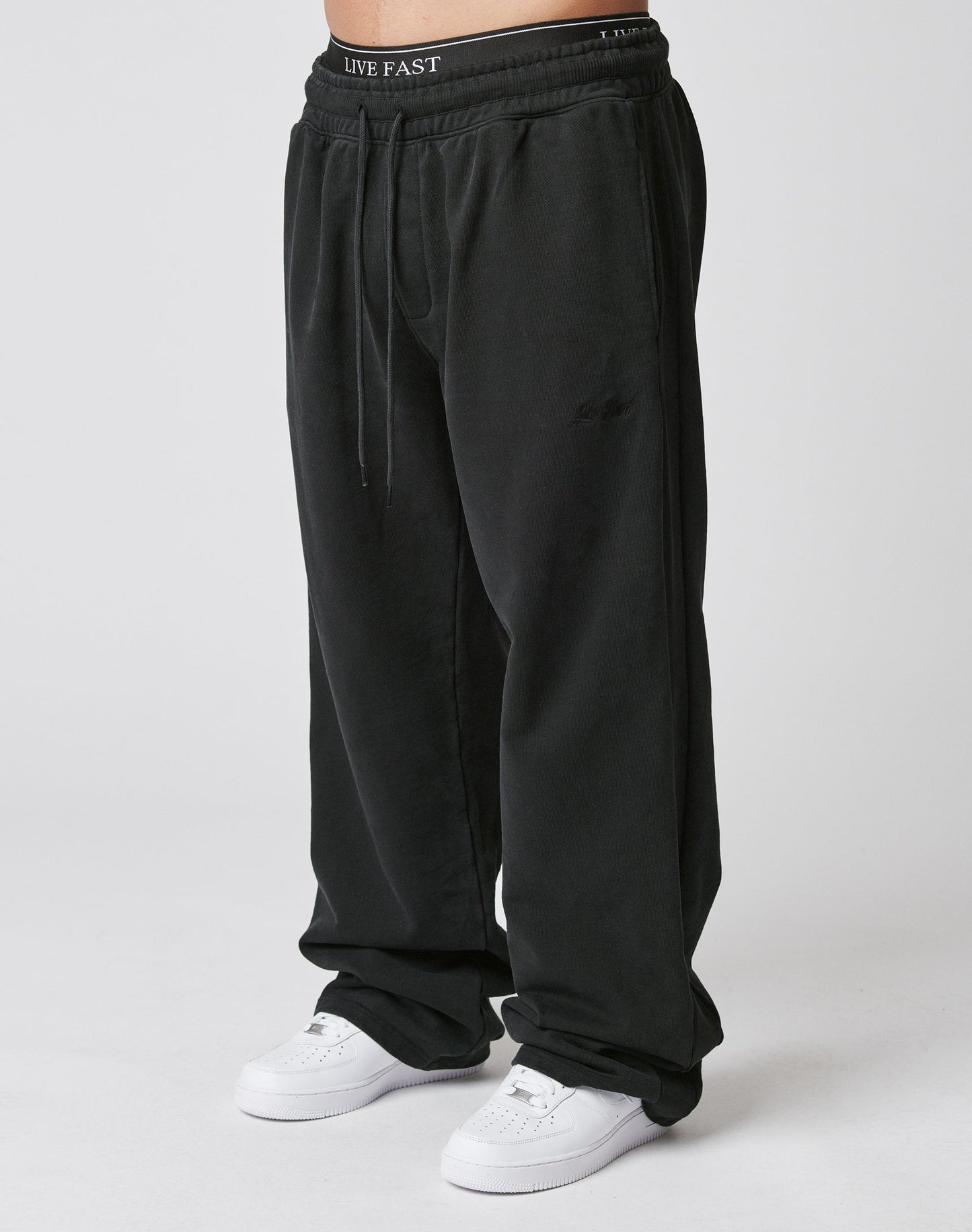 A person in the LIVE FAST DIE YOUNG Basic Wide Jogger—wide fit black sweatpants with an elastic waistband—stands against a plain white background, paired with white sneakers. | model