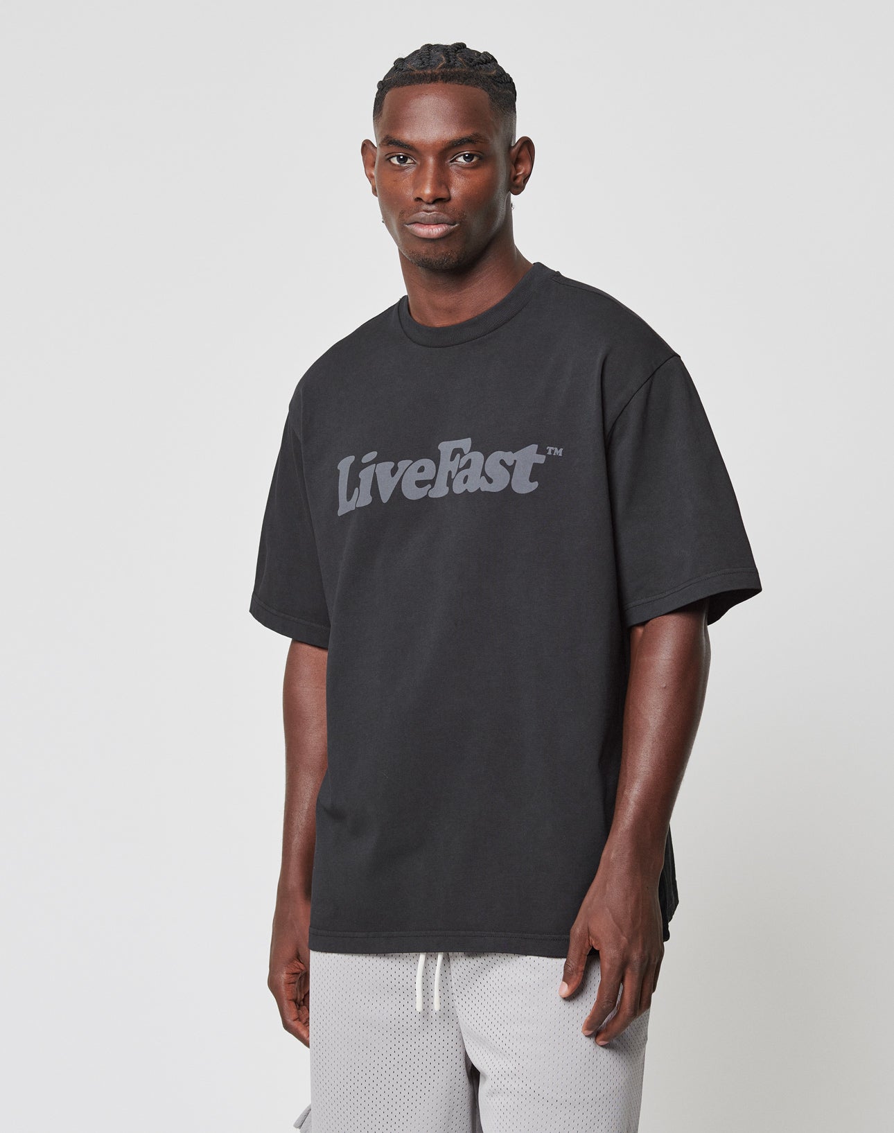 A man wears the LIVE FAST DIE YOUNG Basic Tee—a loose, black, 100% Baumwolle, oversize-fit t-shirt with LiveFast in bold gray on the front—paired with light gray mesh shorts against a plain white background.
