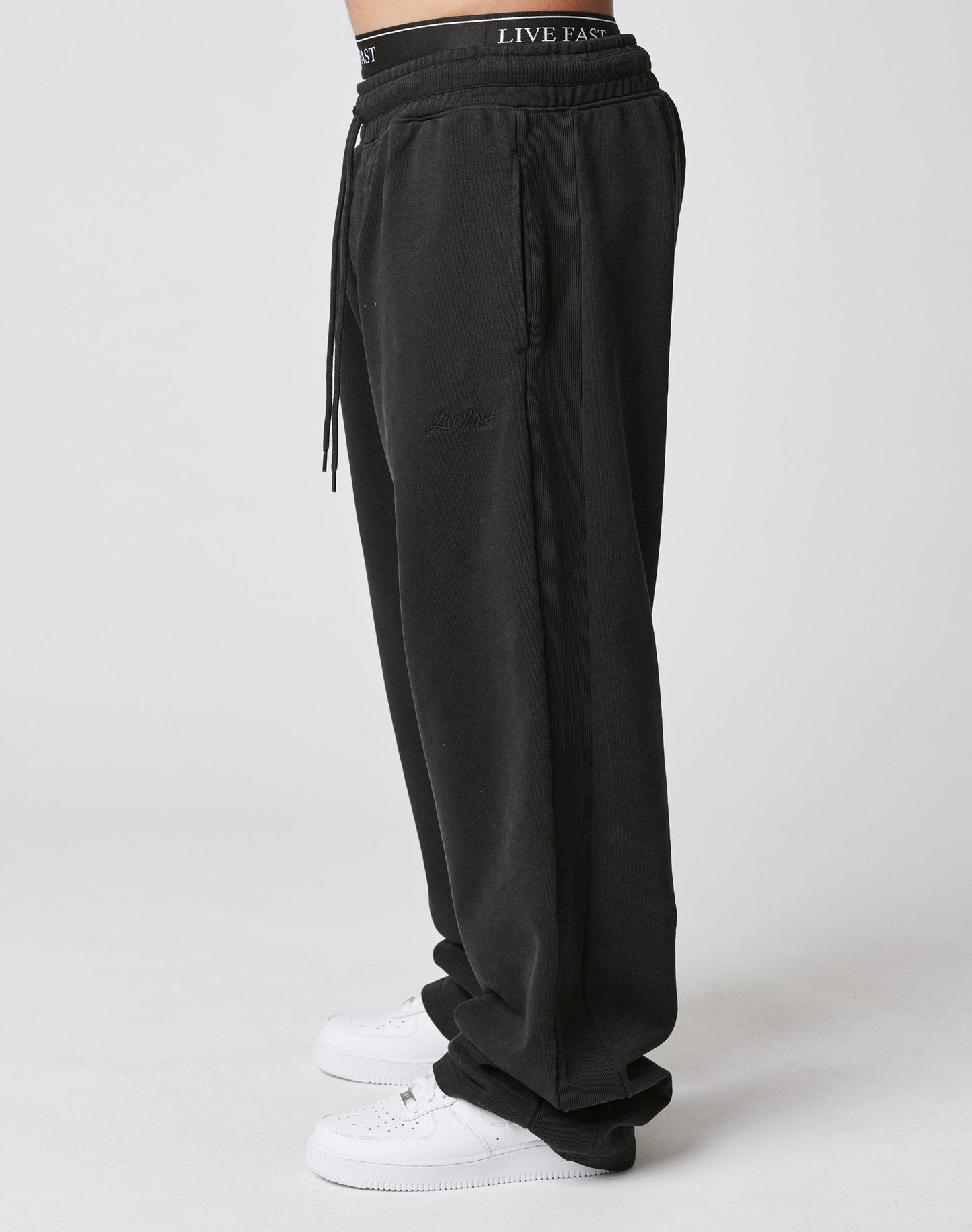 A person wears LIVE FAST DIE YOUNG Basic Wide Jogger, loose-fitting black sweatpants with elastic waistband, hands in pockets, white sneakers, and visible LIVE FAST logo on the underwear. Made from soft Baumwolle. Plain white background.