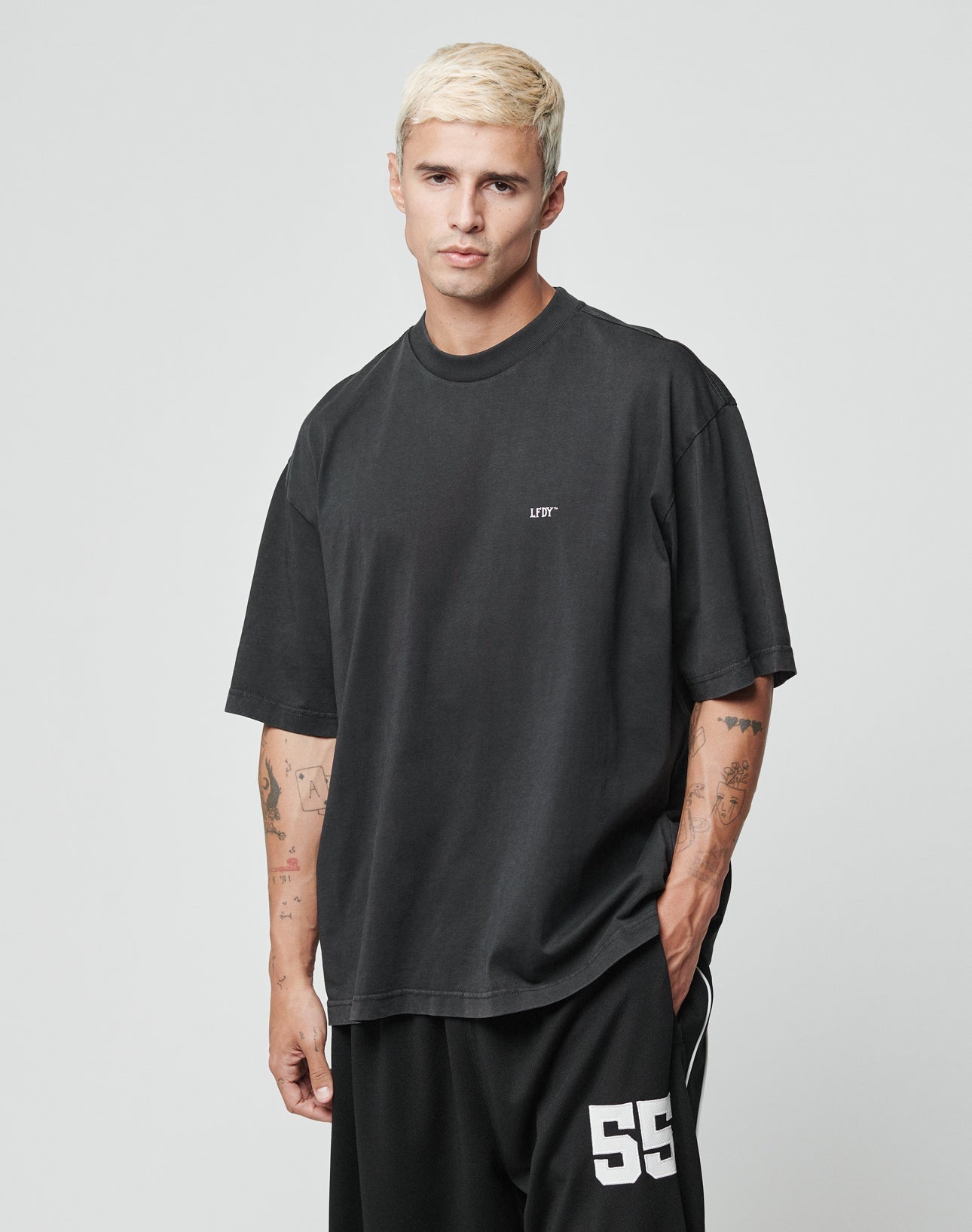 A young man with short bleached blonde hair wears the LIVE FAST DIE YOUNG Everybody Eats Tee, an oversized 100% cotton black T-shirt with LIFT on the chest, paired with black shorts marked 55. He stands against a plain light gray background. Care instructions included.