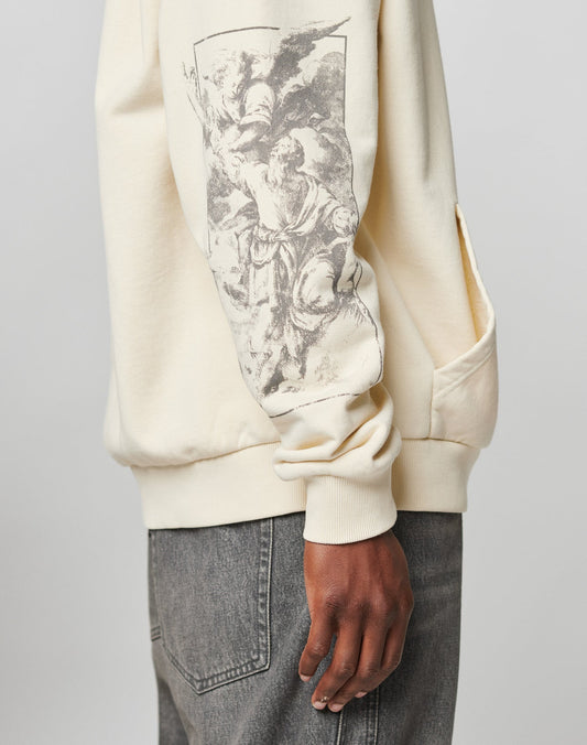 A person wears the LFDY Window Hooded by LIVE FAST DIE YOUNG, an oversized cream sweatshirt with a black-and-white classical art print on the sleeve and subtle logo detail, paired with gray jeans, shown from the side.