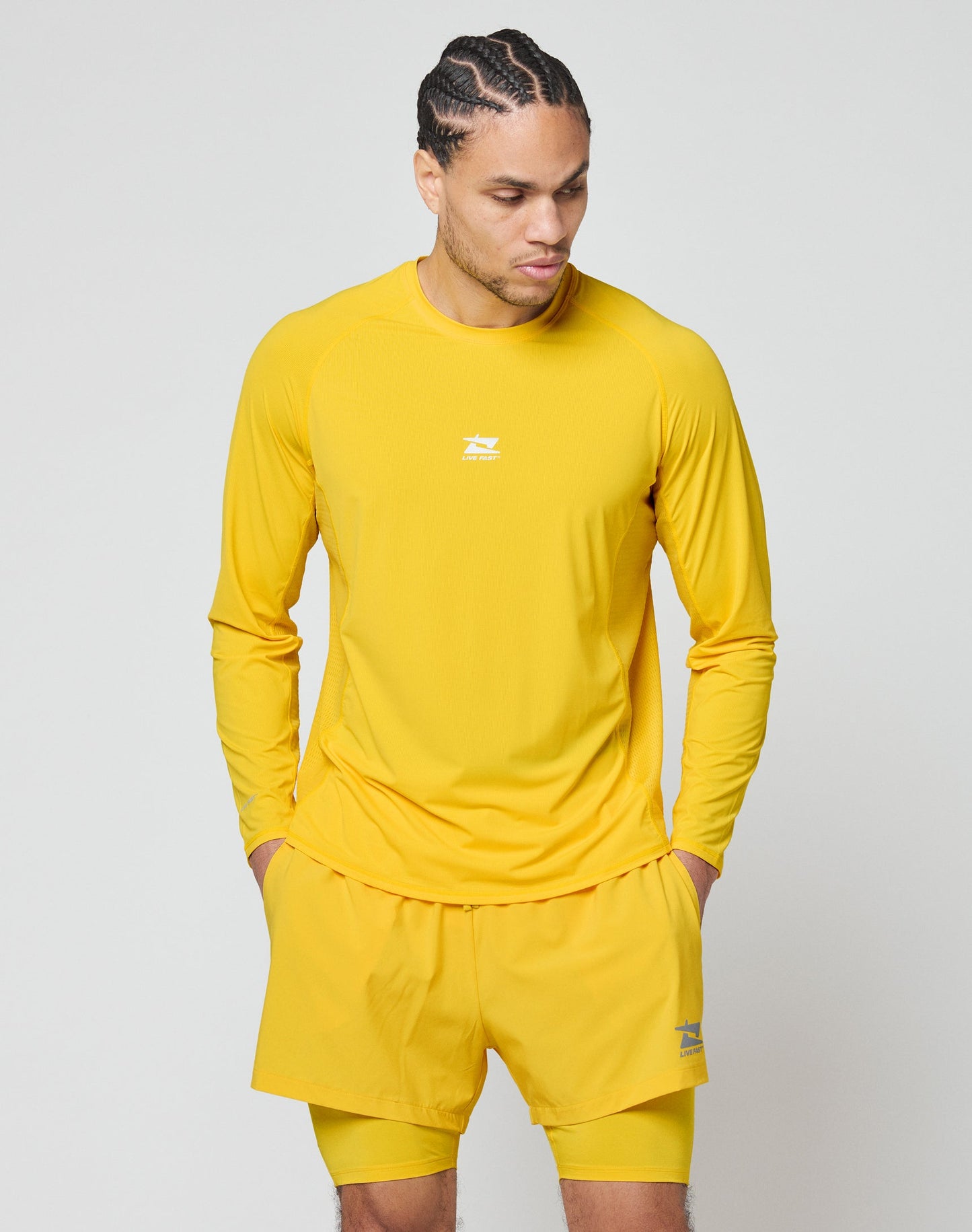 A man wearing the LIVE FAST DIE YOUNG Live Fast Performance Longsleeve and matching yellow shorts with mesh inserts stands with his hands in his pockets, looking down and to the side against a light gray background.