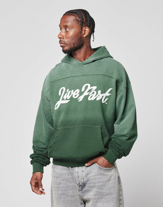 A man with braided hair wears the oversize fit green LFDY Script Hooded by LIVE FAST DIE YOUNG, featuring Live Fast in white script on the front, paired with light-wash distressed jeans, and looks left against a plain background.