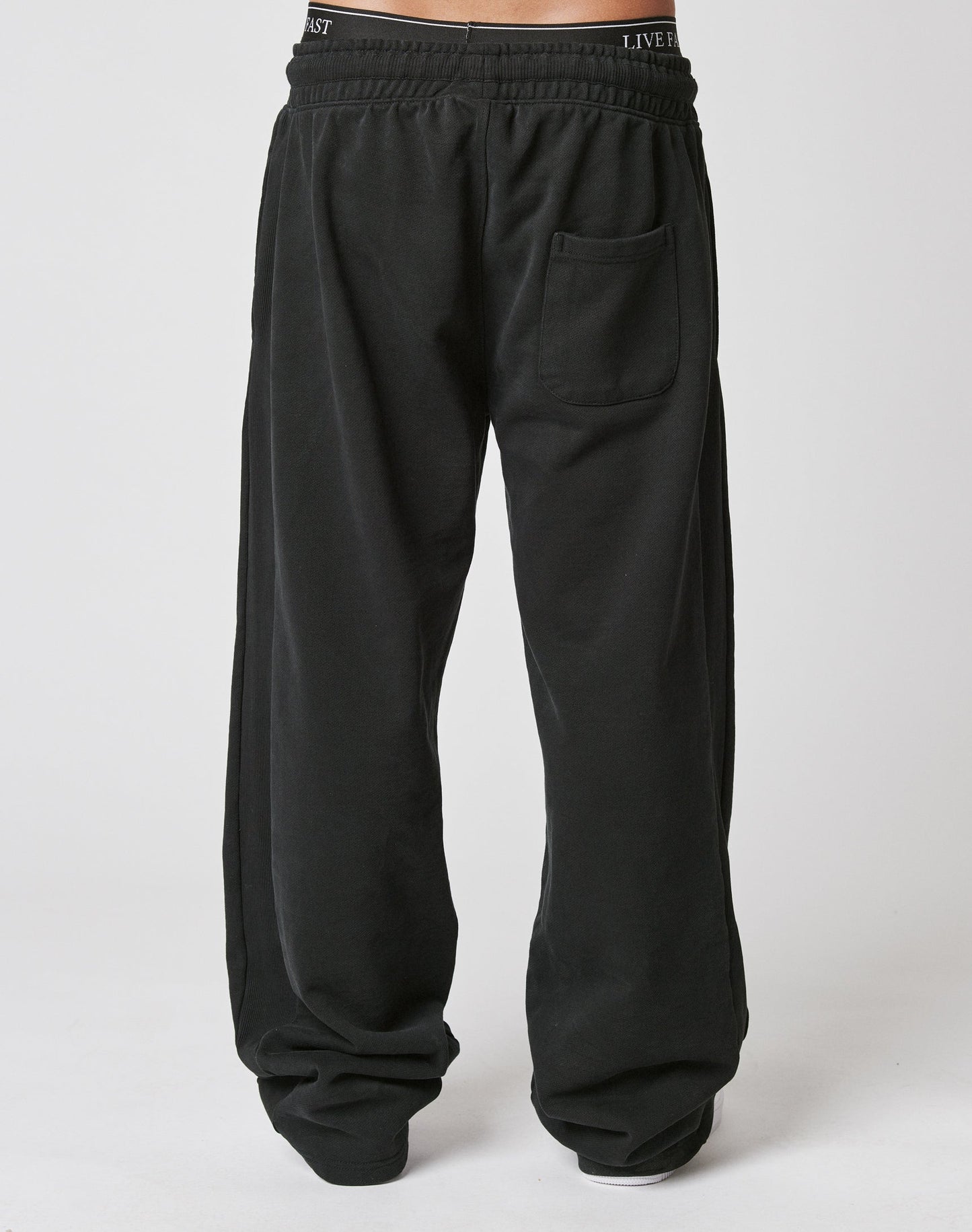 A person wears LIVE FAST DIE YOUNG’s Basic Wide Jogger—loose-fitting black sweatpants with an elastic waistband and wide fit, made from soft cotton. Only their lower half is visible against a plain white background.