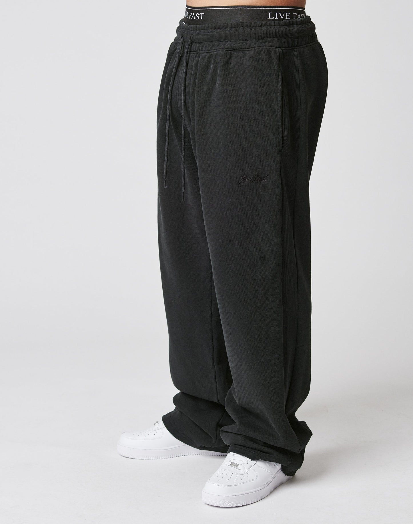 A person wears black Basic Wide Jogger sweatpants from LIVE FAST DIE YOUNG with an elastic waistband featuring the LIVE FAST logo, paired with white sneakers, standing against a plain white background.