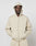 A person in a light beige LIVE FAST DIE YOUNG Basic Script Hooded Zip with the hood up and an oversize fit stands against a white background, hands in the front pockets and wearing a neutral expression.