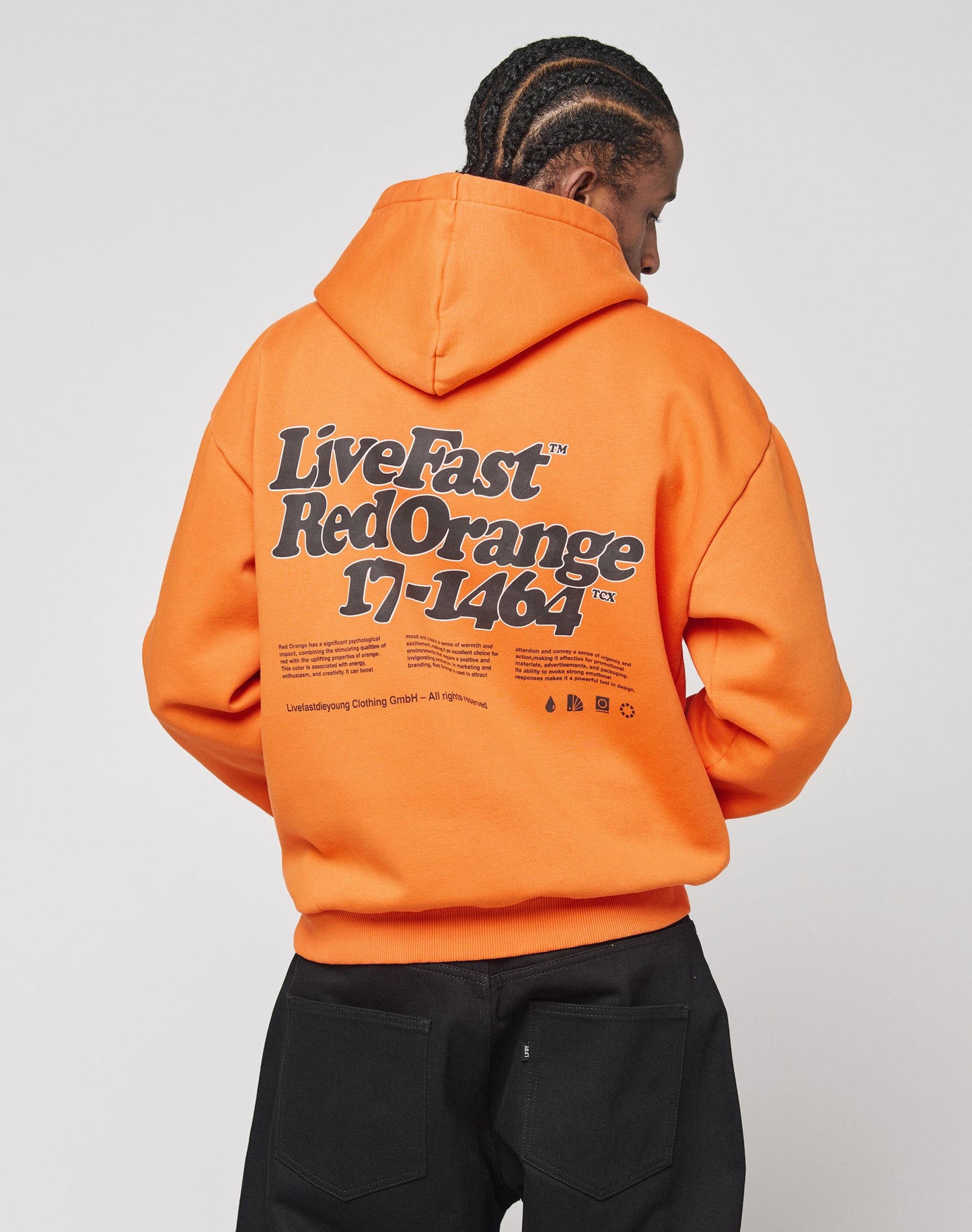 An individual with braided hair faces away, wearing the LIVE FAST DIE YOUNG Basic Orange Hooded made of cotton and polyester, featuring bold front and back “Live Fast Red Orange 17-1464” prints, paired with black pants.