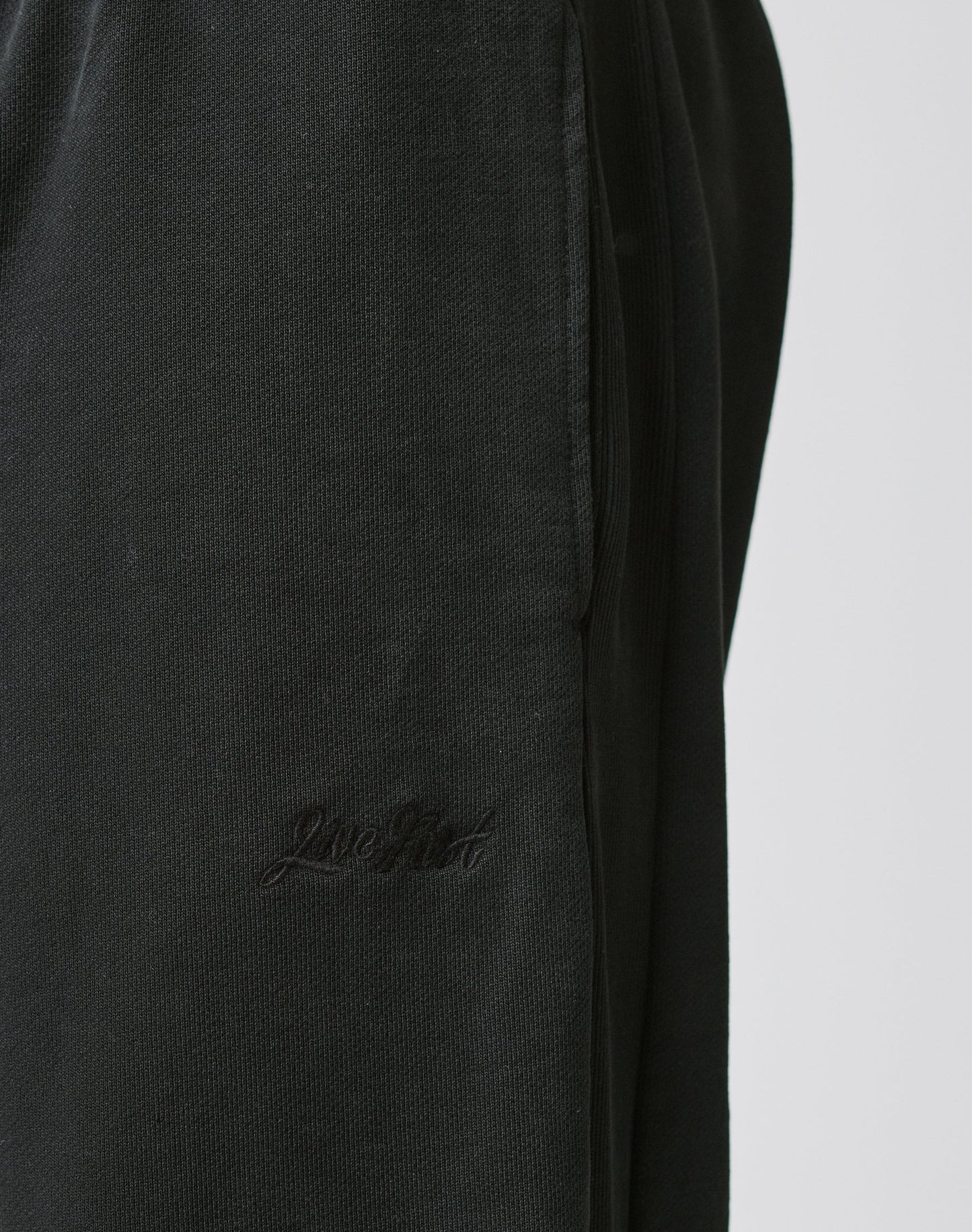 Close-up of Basic Wide Jogger by LIVE FAST DIE YOUNG in black textured cotton, with a wide fit, elastic waistband, and embroidered Live Good logo near the pocket. Shown against a plain, light-colored background.