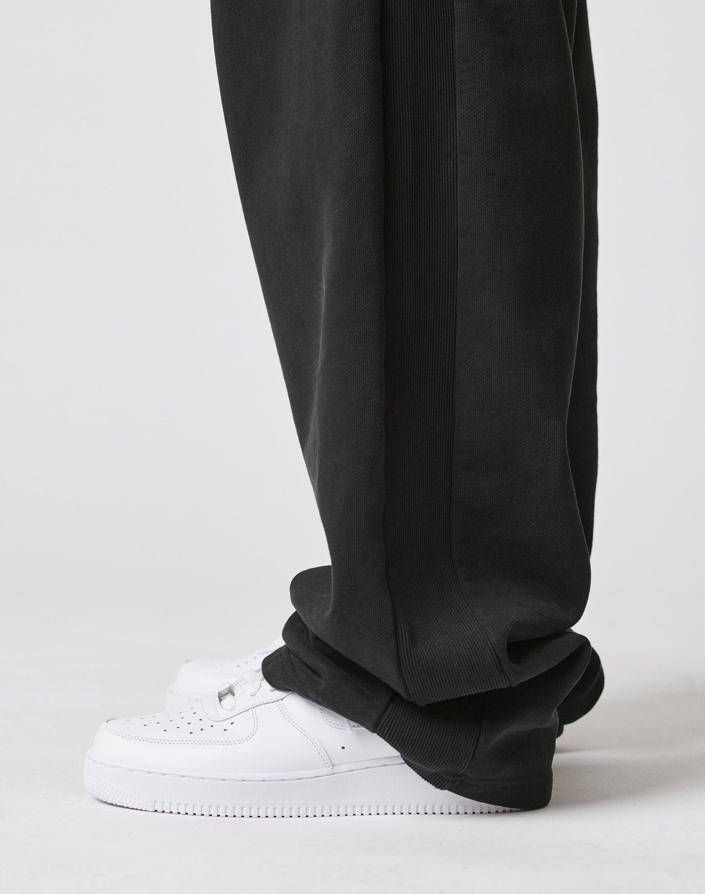 A person is shown from the side at ankle level, wearing LIVE FAST DIE YOUNG Basic Wide Jogger pants in black with a loose fit and elastic waistband, paired with white sneakers on a light-colored floor.