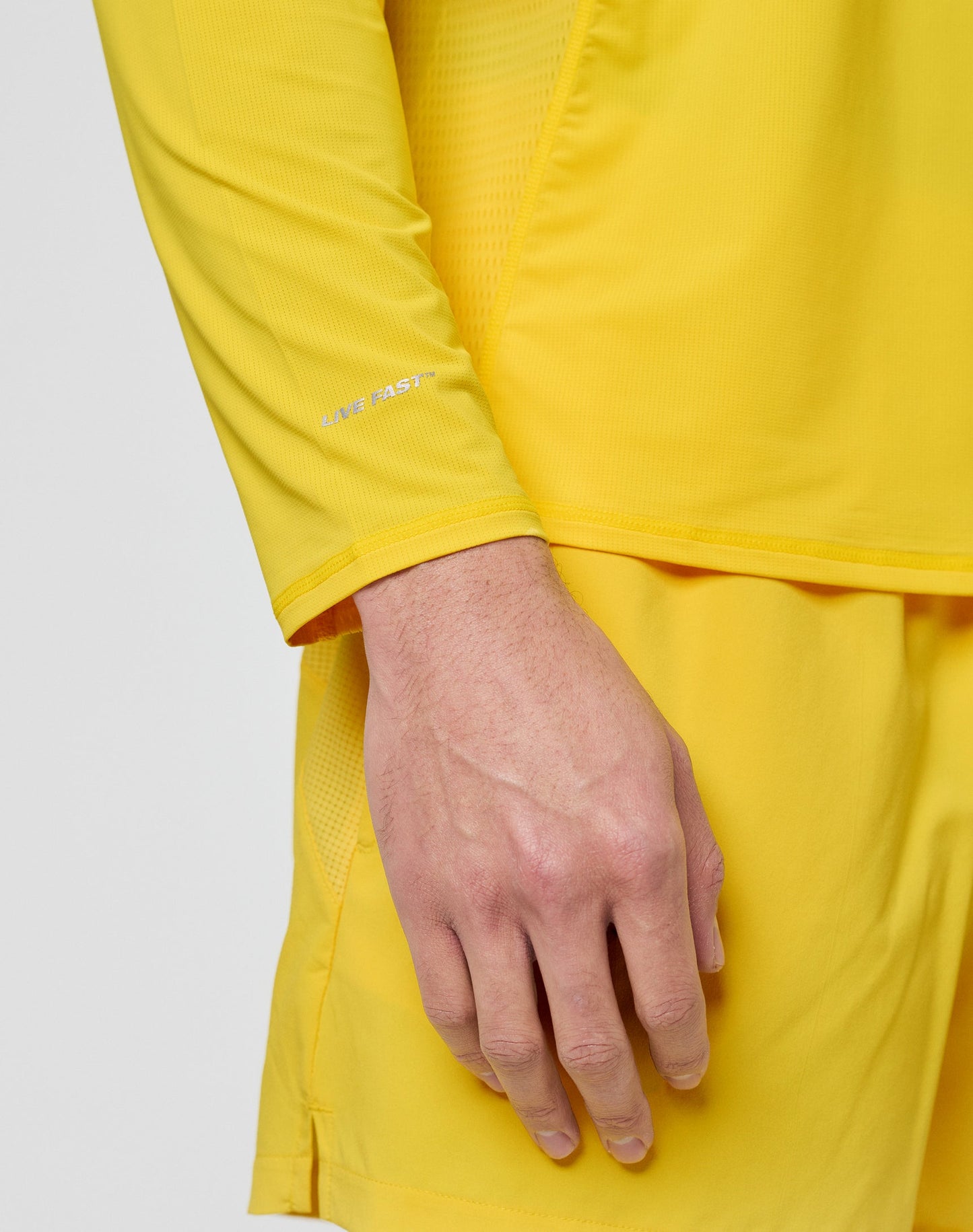 A close-up shows a person’s hand and lower arm wearing the LIVE FAST DIE YOUNG Live Fast Performance Longsleeve in bright yellow with a reflective front print, matching shorts, and LIVE FAST text on the sleeve.