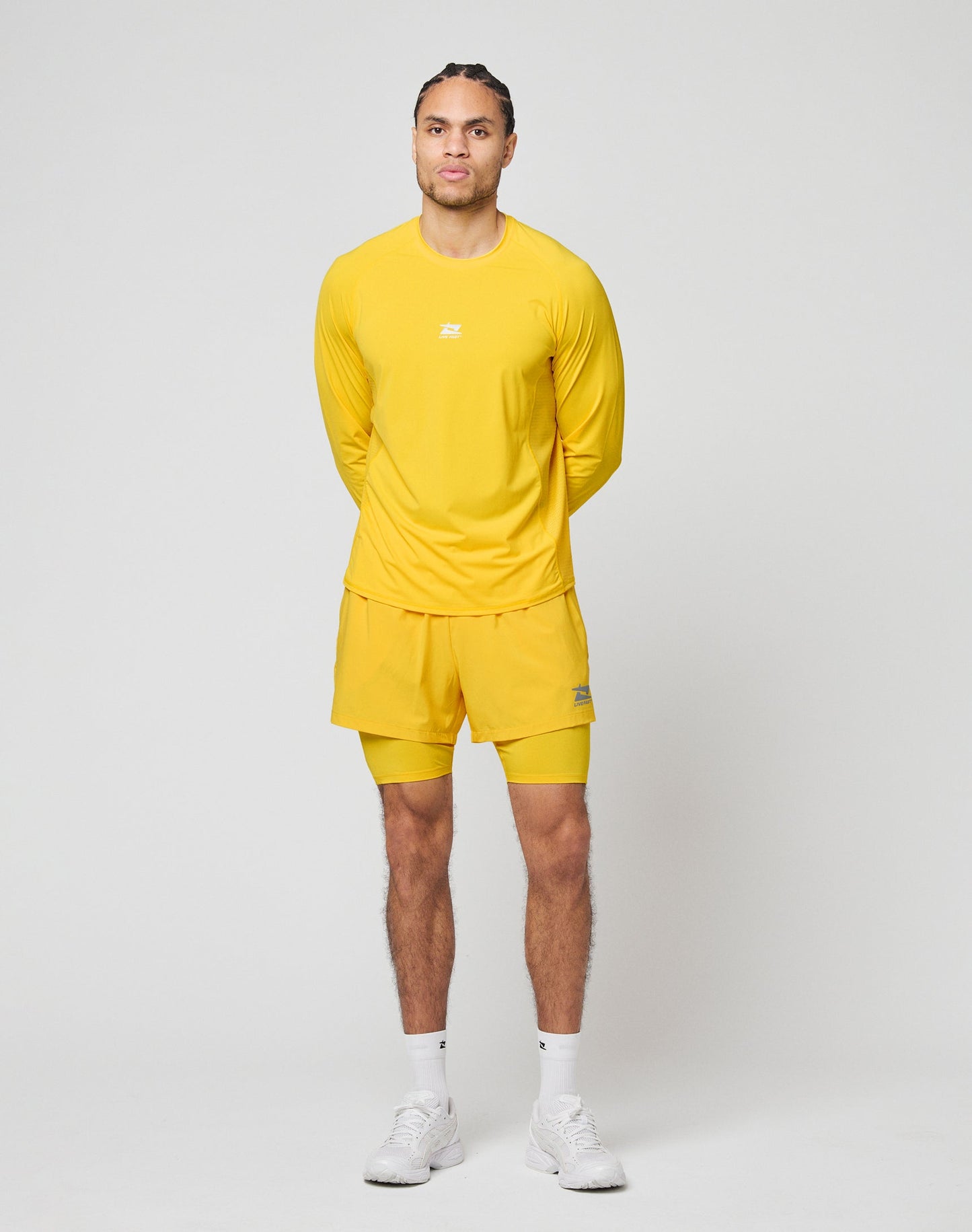 A man stands before a light background in a slim-fit yellow Live Fast Performance Longsleeve by LIVE FAST DIE YOUNG and matching shorts with mesh inserts, paired with white socks and sneakers. His hands are behind his back, expression neutral.