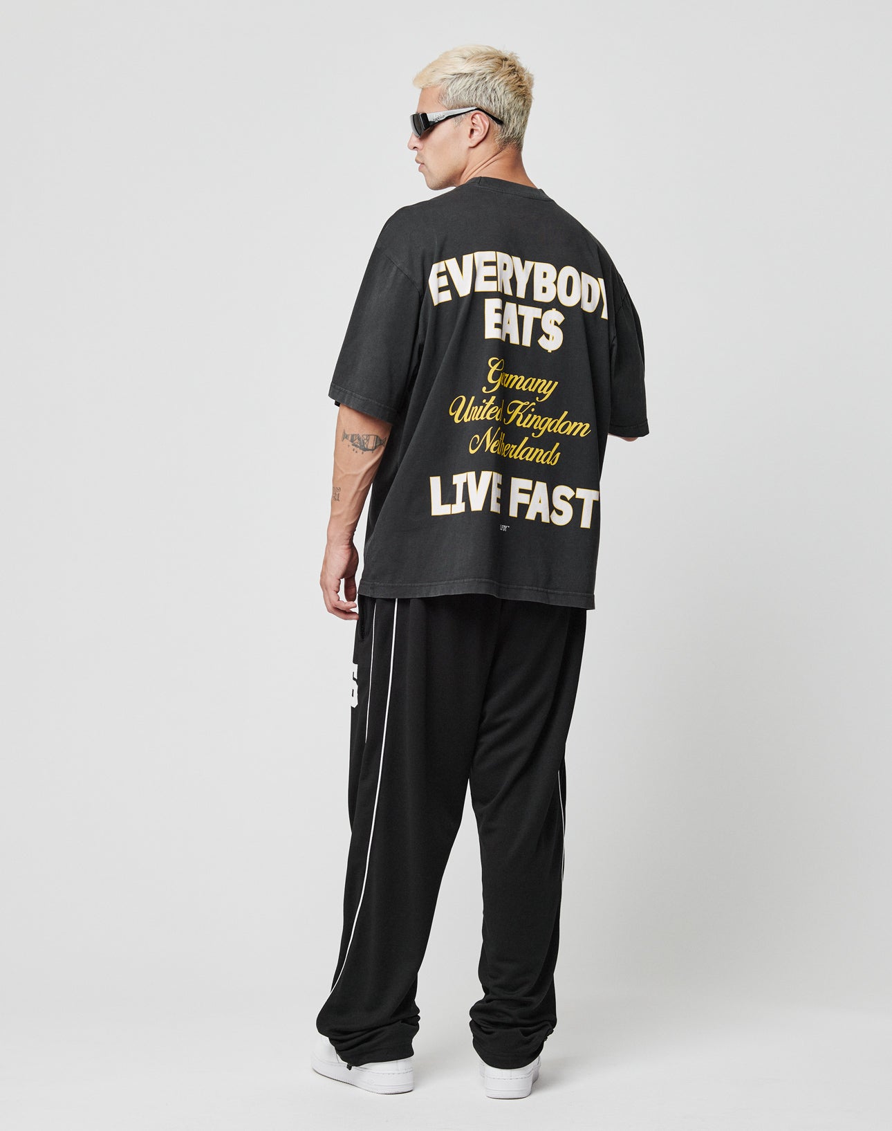 A person with short blond hair, sunglasses, and tattoos stands facing away in the LIVE FAST DIE YOUNG Everybody Eats Tee (black, 100% Baumwolle) featuring “EVERYBODY EATS” and “LIVE FAST” text, black pants, and white shoes against a plain background.