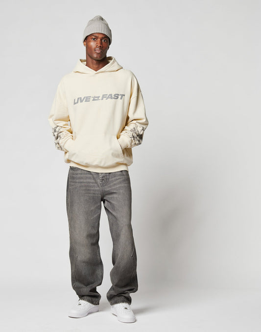 A man wearing a light gray beanie, gray jeans, white sneakers, and the LIVE FAST DIE YOUNG LFDY Window Hooded with reflective LIVE FAST print stands against a plain white background with his hands in his hoodie pocket.