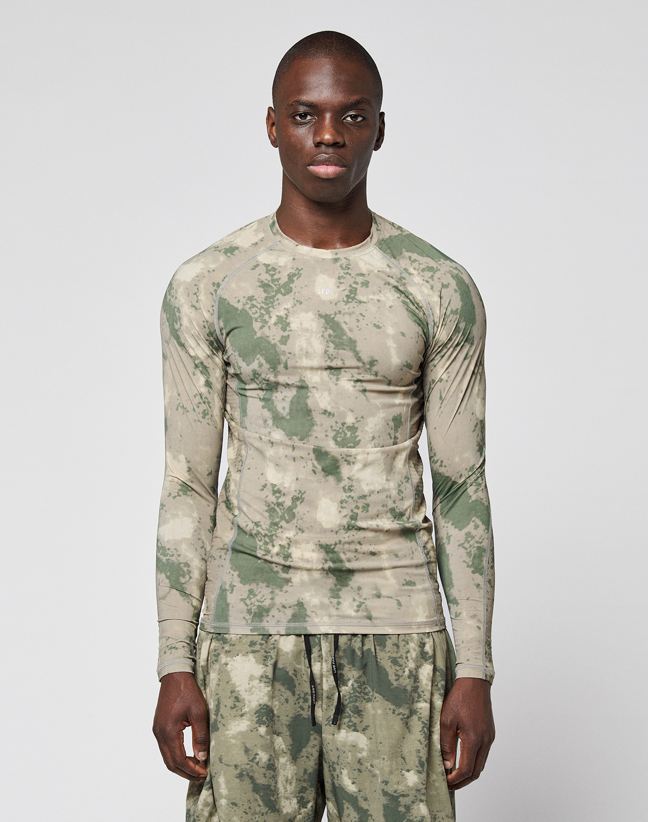 A person wears the LIVE FAST DIE YOUNG LFDY Performance Slim Longsleeve and matching shorts in green and beige camouflage with leichtes Obermaterial, standing against a plain white background. | model