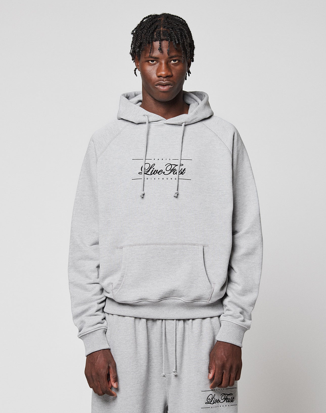 A young man with short braids wears a light gray LFDY Embroidery Hooded by LIVE FAST DIE YOUNG, paired with matching sweatpants. Both pieces feature "Live Fast" and he stands against a plain white background, looking at the camera. | model