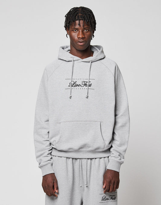 A young man with short braids wears a light gray LFDY Embroidery Hooded by LIVE FAST DIE YOUNG, paired with matching sweatpants. Both pieces feature "Live Fast" and he stands against a plain white background, looking at the camera. | model