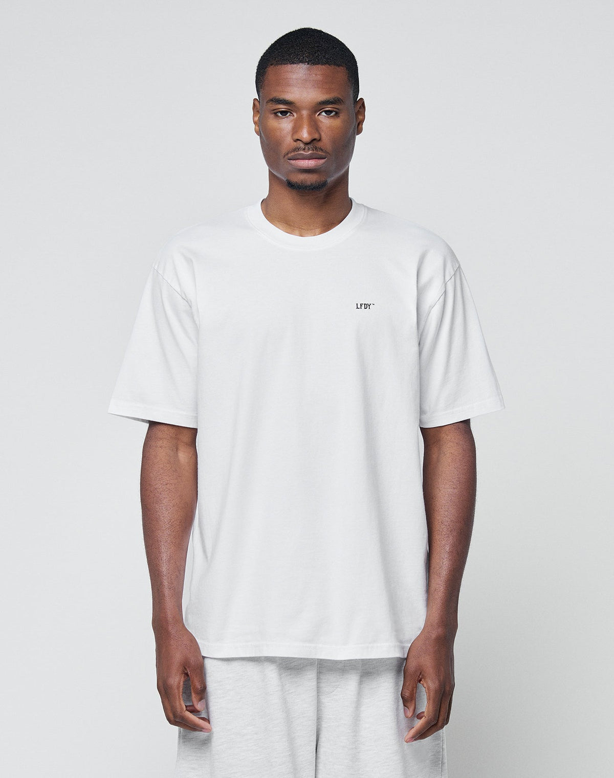 A man stands against a plain light background, wearing LIVE FAST DIE YOUNG’s Basic Chest Tee—a loose-fitting, regular fit white T-shirt with short sleeves and a small LFTP logo print on the left chest—paired with light gray pants. | model
