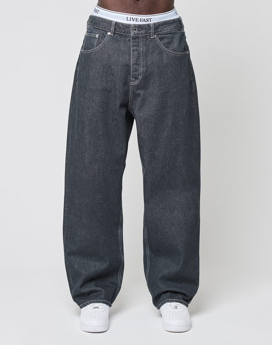 Someone stands against a plain light background, wearing LFDY Baggy Denim by LIVE FAST DIE YOUNG in dark gray 100% Baumwolle, white sneakers, and a visible white waistband with LIVE FAST printed on it.
