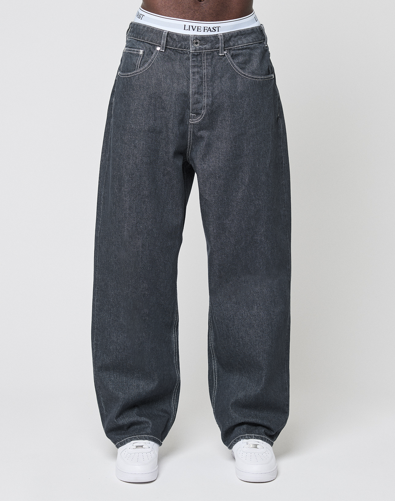 Someone stands against a plain light background, wearing LFDY Baggy Denim by LIVE FAST DIE YOUNG in dark gray 100% Baumwolle, white sneakers, and a visible white waistband with LIVE FAST printed on it.