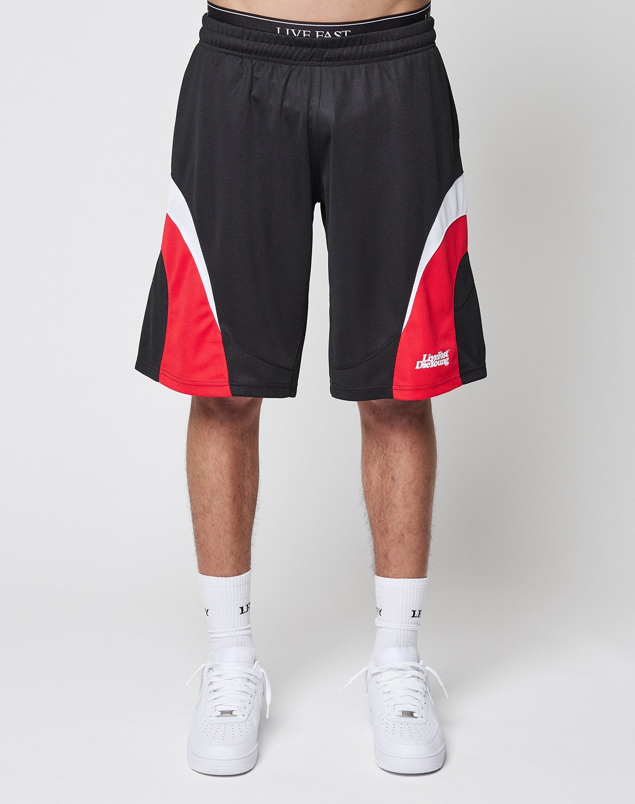 A person is shown from the waist down wearing LIVE FAST DIE YOUNG Basic Trackshorts—black polyester shorts with red and white curved designs—paired with white crew socks and sneakers, set against a plain white background.