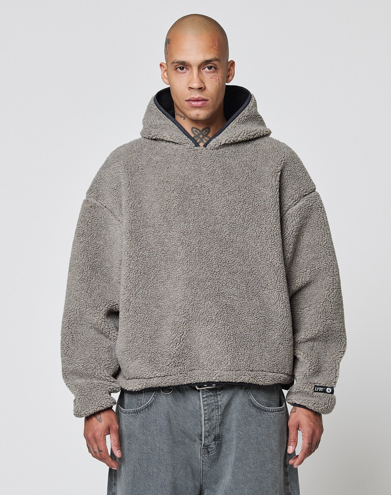 A person with a shaved head wears the LIVE FAST DIE YOUNG LFDY Teddy Hooded in gray, featuring practical Eingrifftaschen, and loose-fitting gray jeans, standing against a plain white background. | model
