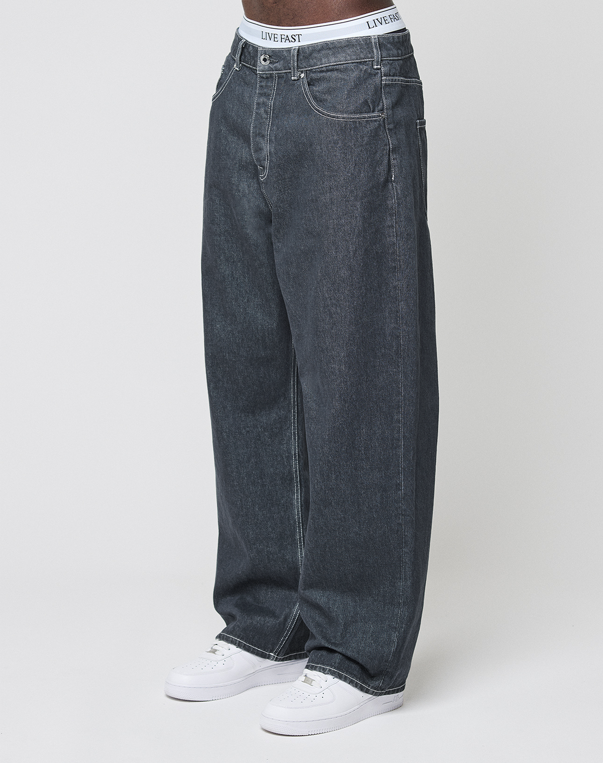A person wears LFDY Baggy Denim by LIVE FAST DIE YOUNG, pairing the dark wash jeans with white sneakers and a visible waistband reading LIVE FAST, against a plain white background. | model