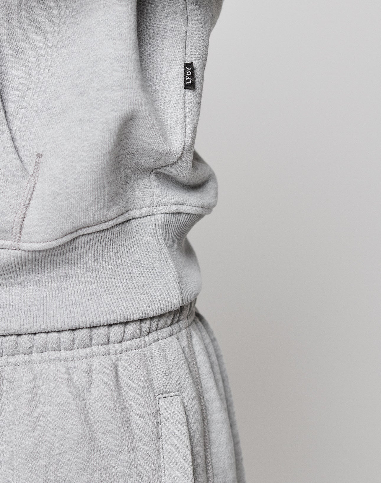Close-up of a person in a light gray LFDY Embroidery Hooded by LIVE FAST DIE YOUNG with raglan sleeves and matching sweatpants. A small black label with white text is on the waistband. The fabric looks soft and comfortable.