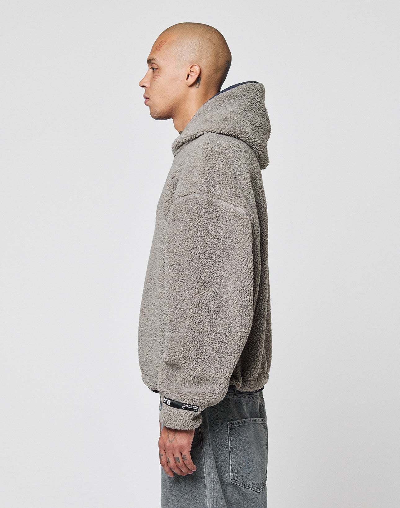 A person with a shaved head stands in profile against a white background, wearing a gray, textured, oversize fit LIVE FAST DIE YOUNG LFDY Teddy Hooded jacket with Eingrifftaschen and faded gray jeans.