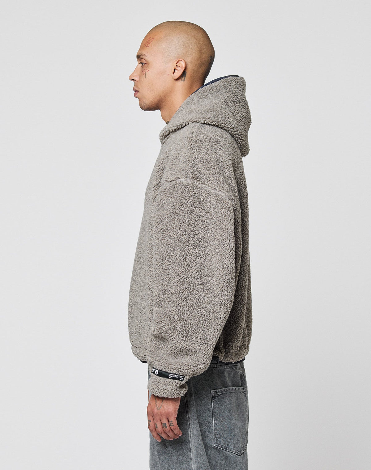 A person with a shaved head stands in profile against a white background, wearing a gray, textured, oversize fit LIVE FAST DIE YOUNG LFDY Teddy Hooded jacket with Eingrifftaschen and faded gray jeans.