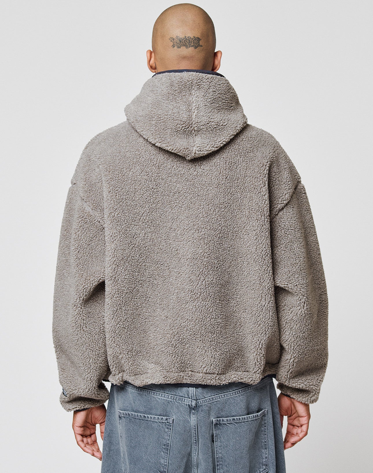 A person with a shaved head and a tattoo wears the LIVE FAST DIE YOUNG LFDY Teddy Hooded jacket, featuring an oversized fit and Eingrifftaschen, paired with denim pants, standing with their back to a plain white background.