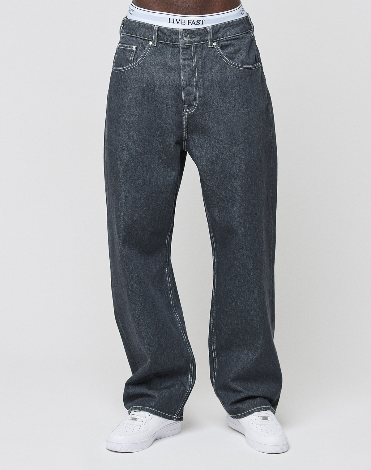 A person wears LIVE FAST DIE YOUNG’s LFDY Baggy Denim jeans, white sneakers, and white underwear with a visible LIVE FAST waistband, shown from waist to feet against a plain light background.