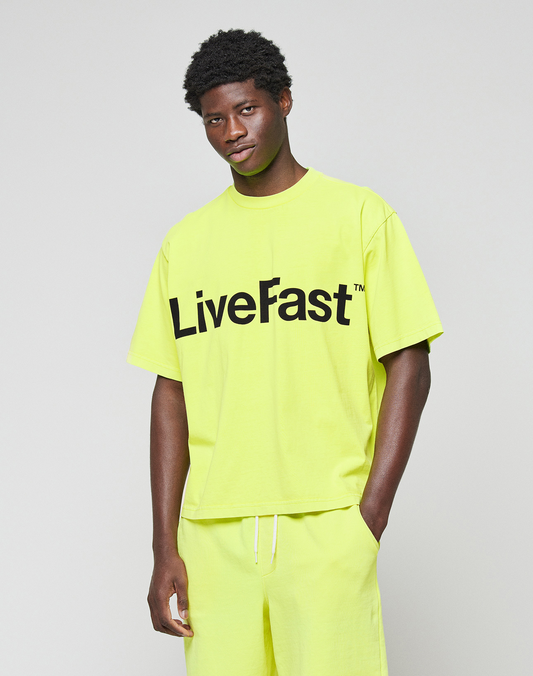 A man stands against a plain background wearing the LIVE FAST DIE YOUNG Basic OS Livefast Tee, an oversized bright yellow T-shirt made from 100% Baumwolle with "LiveFast" printed in large black letters on the front.